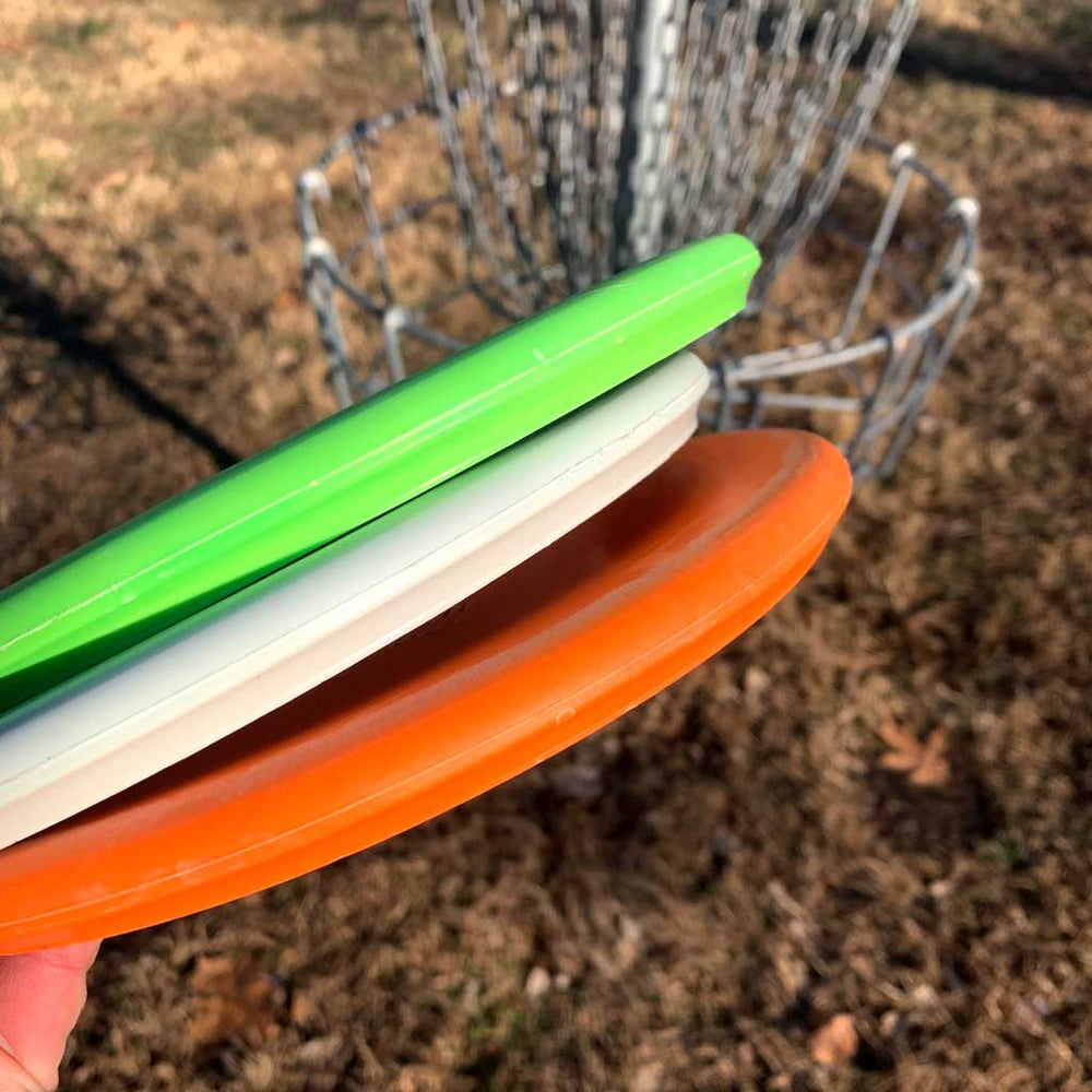 A hand holds three Chief OS - Suregrip disc golf discs—green, white, and orange—ideal for overstable forehand drives in front of a disc golf basket on the grass.