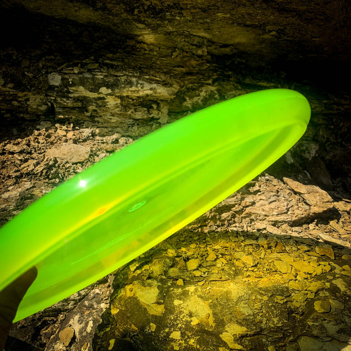 A person's hand holds a bright green Chief - Diamond putt & approach disc with a beaded rim in front of a rocky, sunlit background.