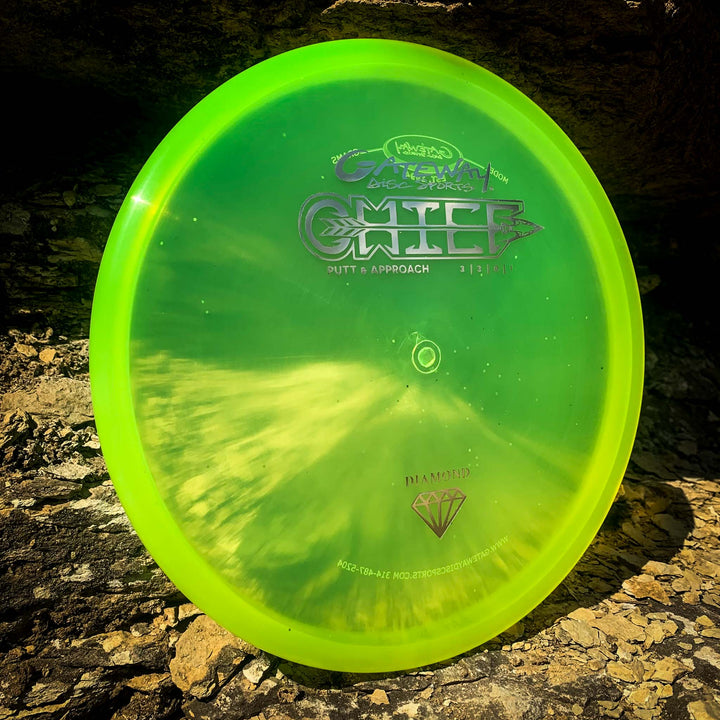 A bright green and yellow Chief - Diamond disc golf putter with a beaded rim rests against rocks in sunlight.