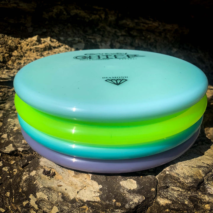 A stack of four colorful disc golf discs—blue, green, yellow, and purple—including the beaded rim Putt & Approach putter Chief - Diamond—rests on a rocky surface in sunlight.