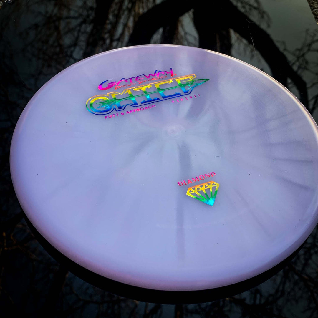 A lavender Gateway Chief - Diamond putter with a beaded rim, iridescent lettering, and a diamond logo sits on a reflective dark surface with tree branches in the background.