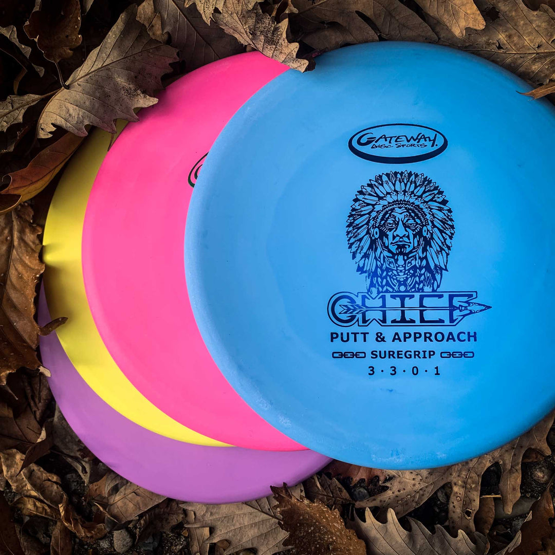 Three colorful Chief - SureGrip disc golf discs—blue, pink, and yellow—rest on brown fallen leaves. The blue disc shows a beaded rim for control, the text "Chief Putt & Approach," and a Native American chief illustration.
