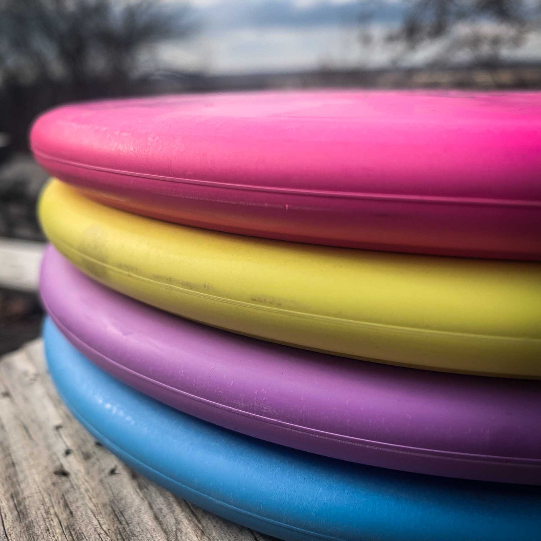 A close-up of four stacked Chief - SureGrip discs for Putt & Approach, featuring beaded rims in pink, yellow, purple, and blue on a wooden surface outdoors—ideal for extra control in your game.