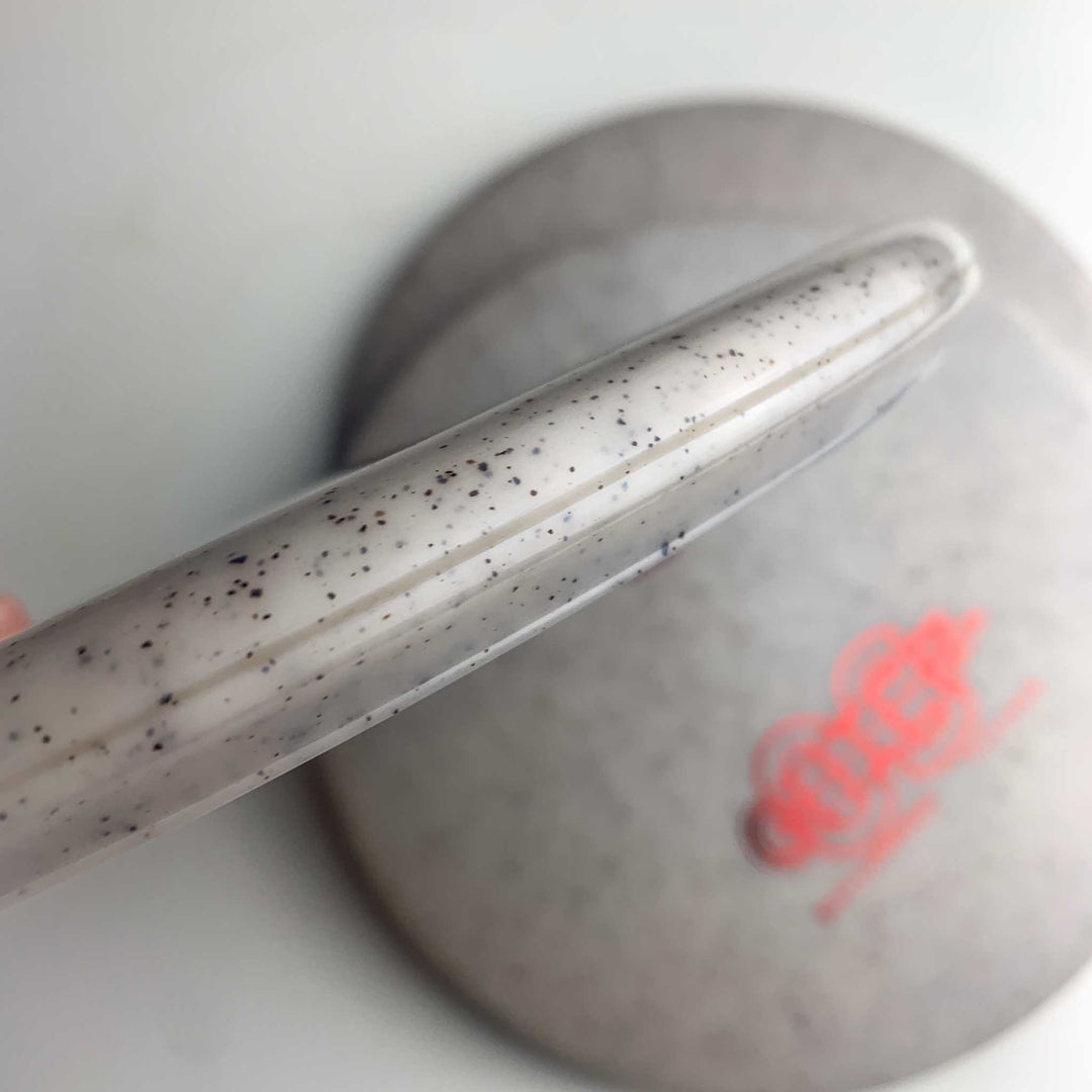 Close-up of a grey, speckled Chief OS - Diamond Hemp disc featuring a slightly translucent rim; its overstable design is perfect for precise hyzer line forehand drives. Another disc with red text appears in the background on a white surface.