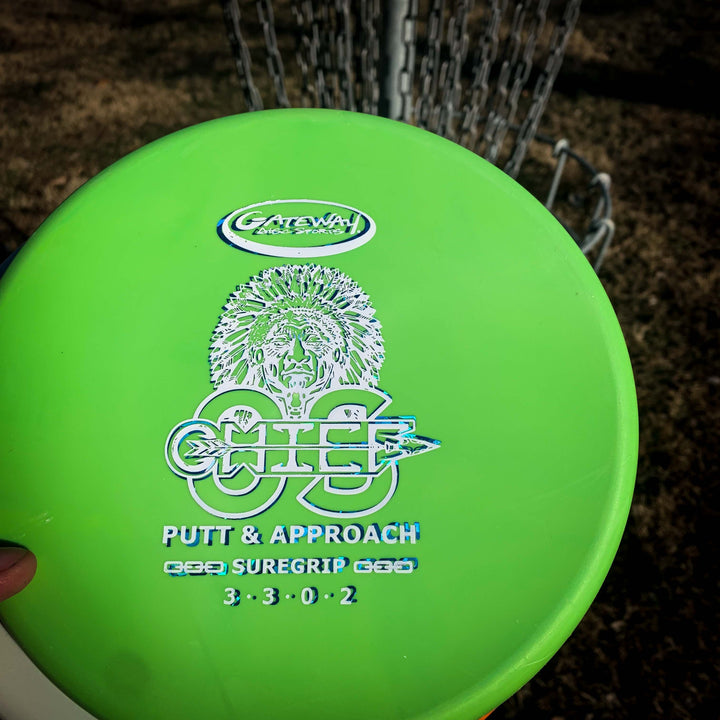 A close-up of the green, overstable Gateway Chief OS - Suregrip disc golf putter, labeled "PUTT & APPROACH SUREGRIP 3·3·0·2," held near a basket—ideal for hyzer shots and consistent control.