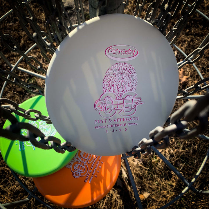 Three Chief OS - Suregrip disc golf discs—two green, one white—sit in a metal basket, ideal for overstable forehand drives. Sunlight casts shadows on the dry grass in the background.