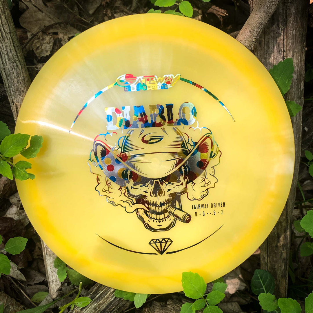 A yellow Diablo - Diamond overstable fairway driver disc featuring a colorful skull with cowboy hat design and —Fairway Driver 9.5.5.-1—� label rests among leaves and branches, ideal for accurate landing shots.