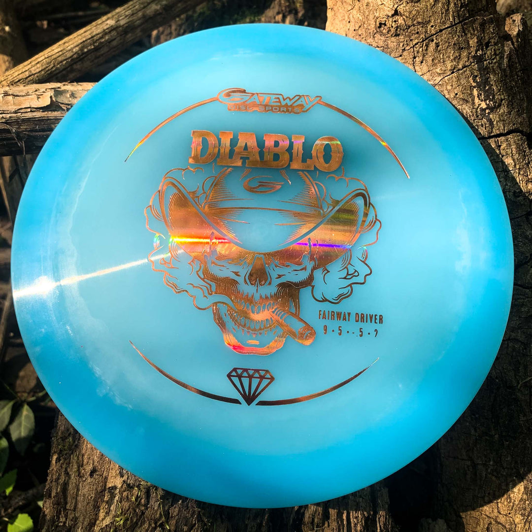 A blue Diablo - Diamond overstable fairway driver disc golf disc with a holographic skull design and text rests on a tree trunk.