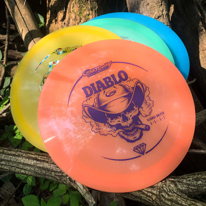 Three brightly colored disc golf discs, including an orange Diablo - Diamond control driver with a skull graphic, are resting on tree branches outdoors.