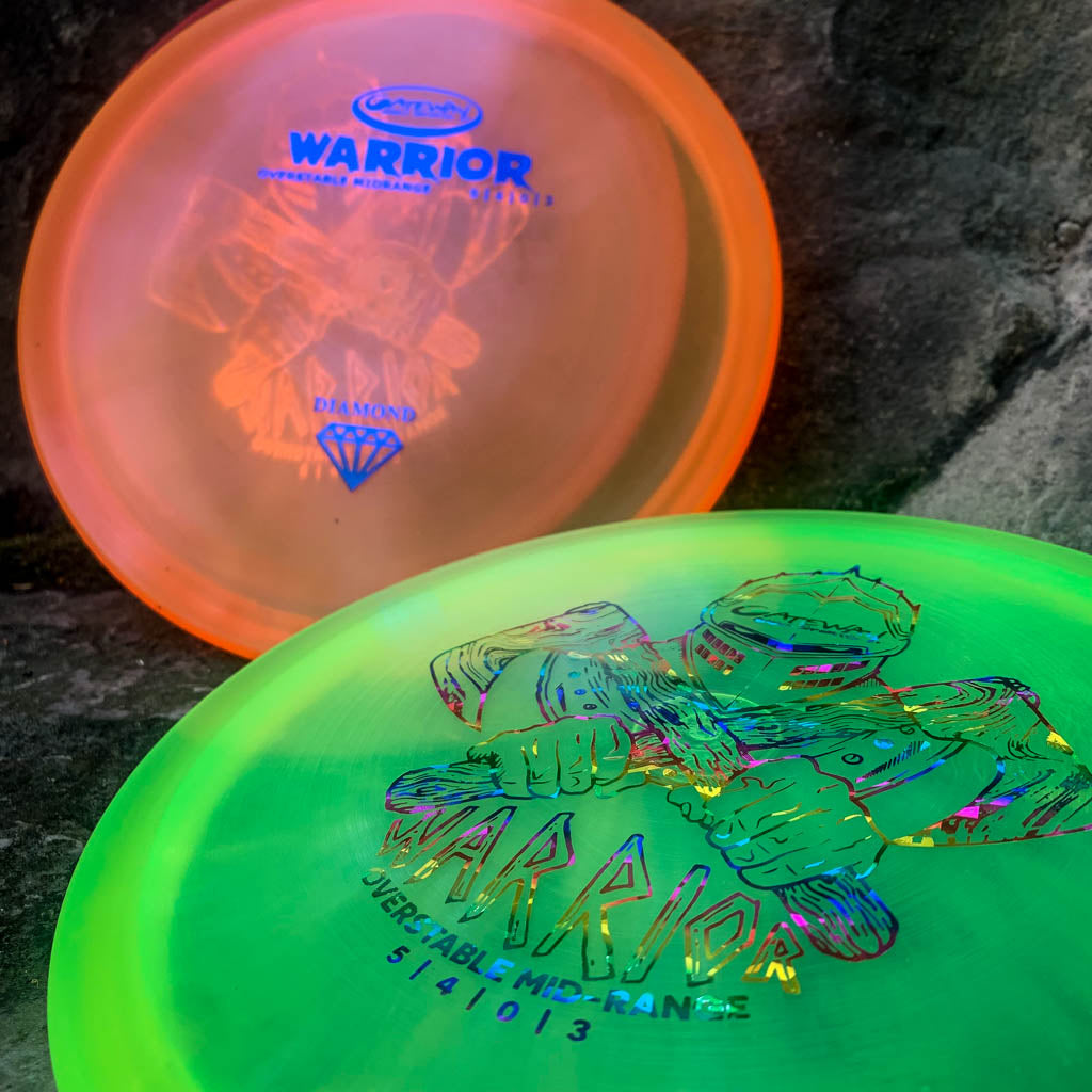 Two vibrant mid-range disc golf discs, one orange and one green, with "Warrior - Diamond" branding and designs, are displayed on a dark stone surface. The Warrior - Diamond is known for its dependable, overstable flight.