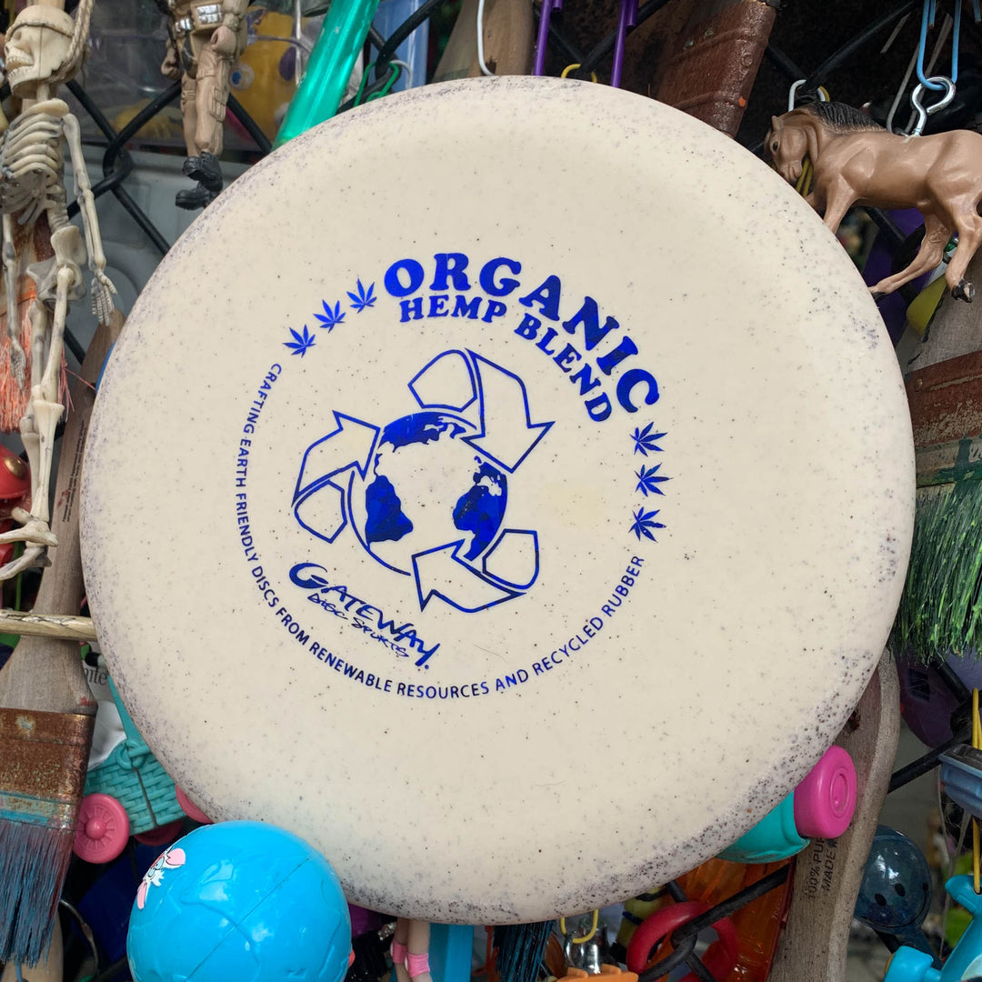 A white "Wizard - SSS - Hemp" disc golf disc featuring an "Organic Hemp Blend" label, blue recycle symbol, and earth graphic; ideal for mid-range drives, pictured with assorted toys and objects in the background.