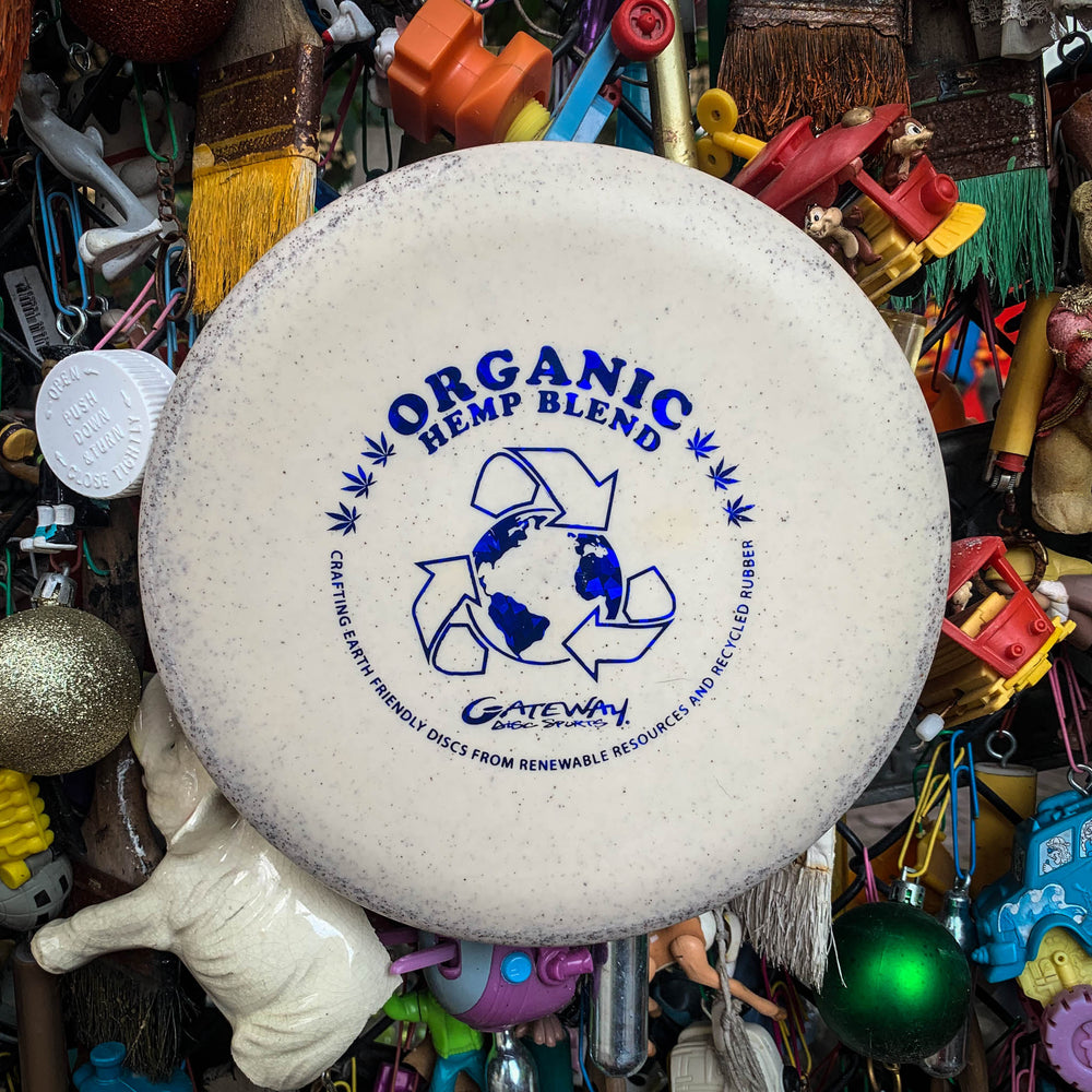 A white Voodoo - Organic Hemp Super Soft (H-SS) disc golf putter with a recycling symbol is shown against a backdrop of colorful objects and toys.