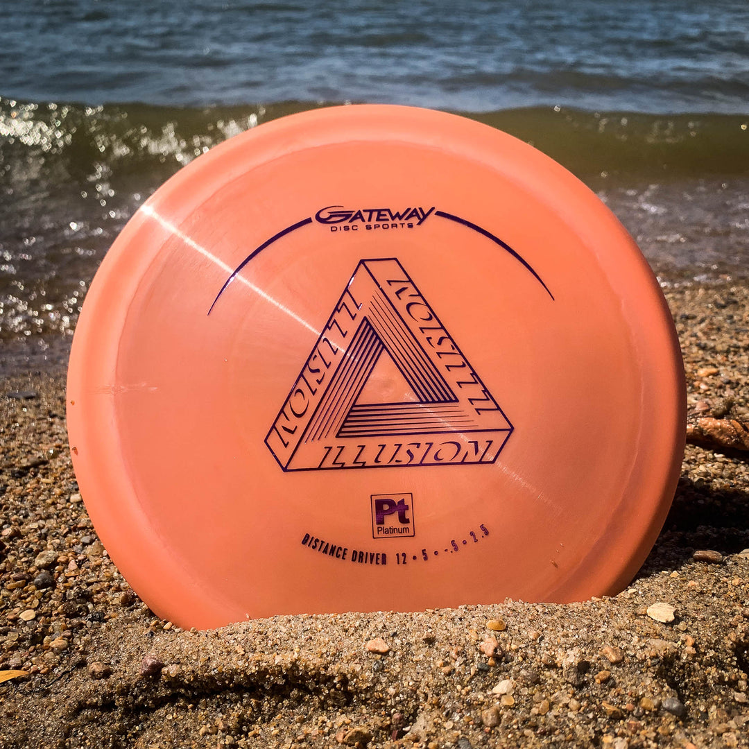 A Platinum Illusion disc golf disc, ideal for powerful distance drives and sidearms, rests partially buried in sand near the water's edge.