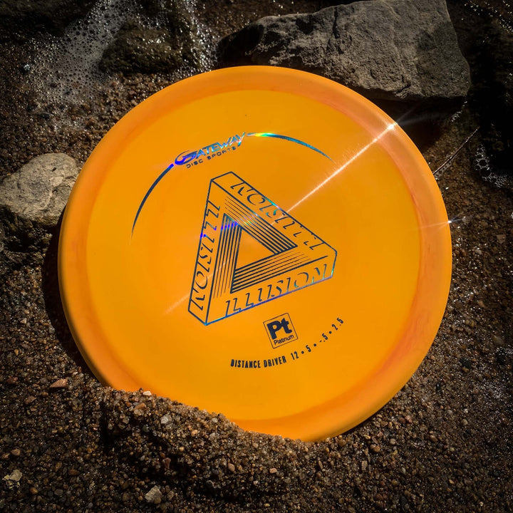 The Illusion - Platinum disc golf driver, partially buried in sand near rocks with sunlight glinting off its surface—ideal for distance throws or sidearms on tough courses.