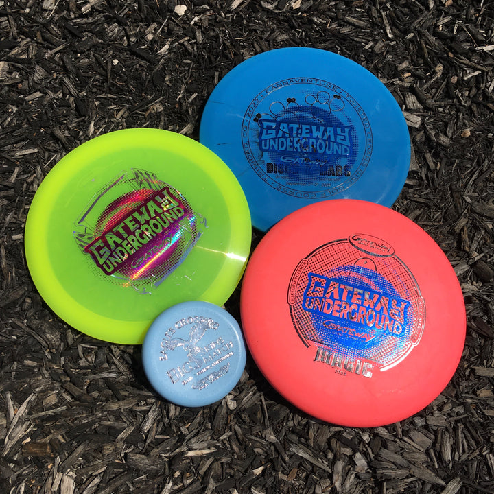 Three discs from the Factory 2nd 3-Disc Starter Pack—yellow, blue, and red—plus a small light blue SureGrip putter are arranged on dark wood chips, each showing distinct Gateway or Blizzard logos.
