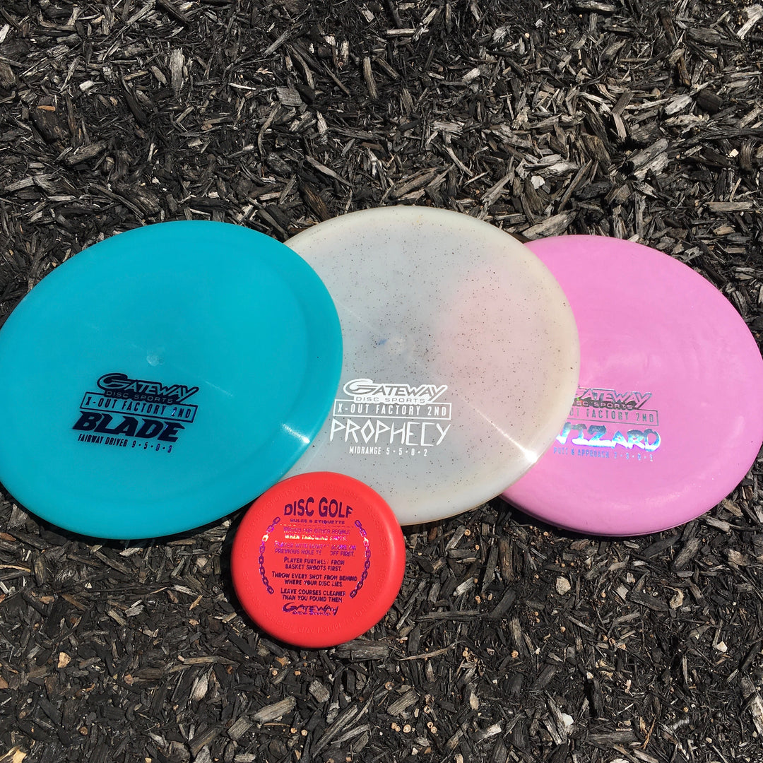 Four disc golf discs in blue, white, pink, and red are displayed on mulch, including a Premium driver and SureGrip putter from the Factory 2nd 3-Disc Starter Pack.