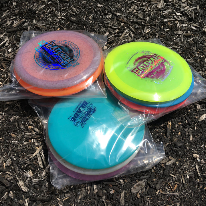Three stacks of Factory 2nd 3-Disc Starter Packs, each with SureGrip putters and premium drivers in plastic bags, are arranged on dark wood mulch outdoors.