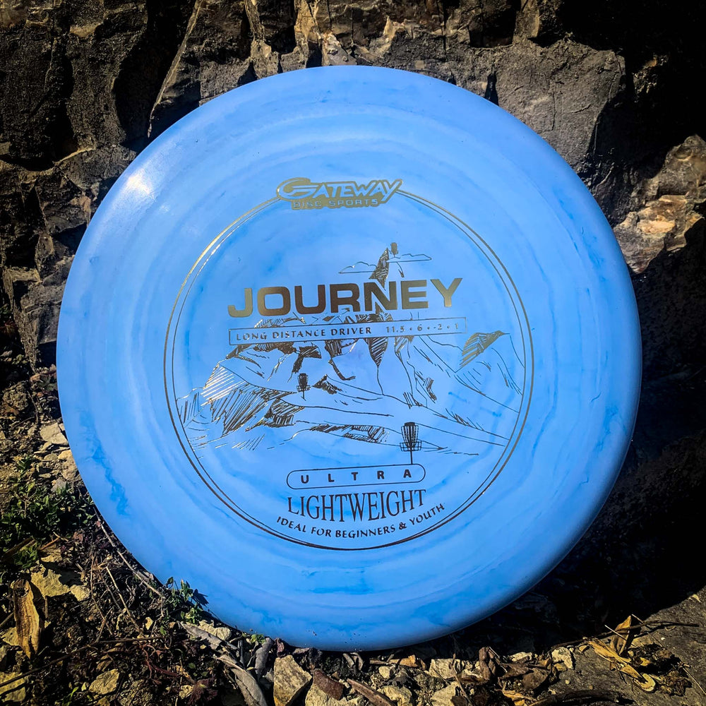 A blue Journey - Ultra Light Weight disc golf disc with mountain and basket graphics leans on rocks. This lightweight distance driver is perfect for beginners and youth seeking easy, controllable flight on the course.