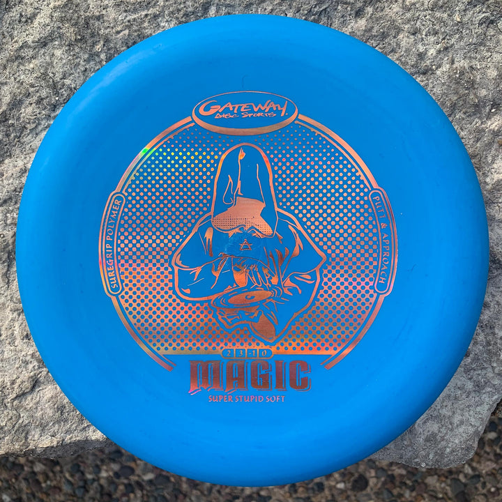 A blue Gateway Magic - Suregrip - SSS putter with a hooded figure and "Magic" & "Super Stupid Soft" text sits on a rock. This straight-flying, easy-to-control disc is ideal for precise putting.