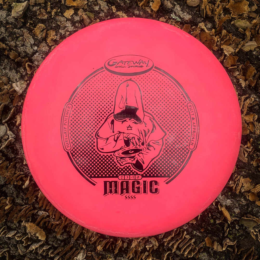 A pink Gateway Magic - Suregrip - SSSS putter with a hooded figure graphic lies on wood chips and leaves. This straight-flying, easy-to-control disc suits both beginners and experienced putters.