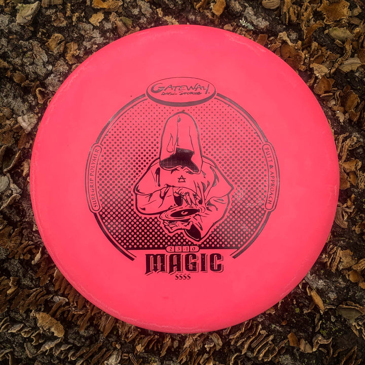 A pink Gateway Magic - Suregrip - SSSS putter with a hooded figure graphic lies on wood chips and leaves. This straight-flying, easy-to-control disc suits both beginners and experienced putters.