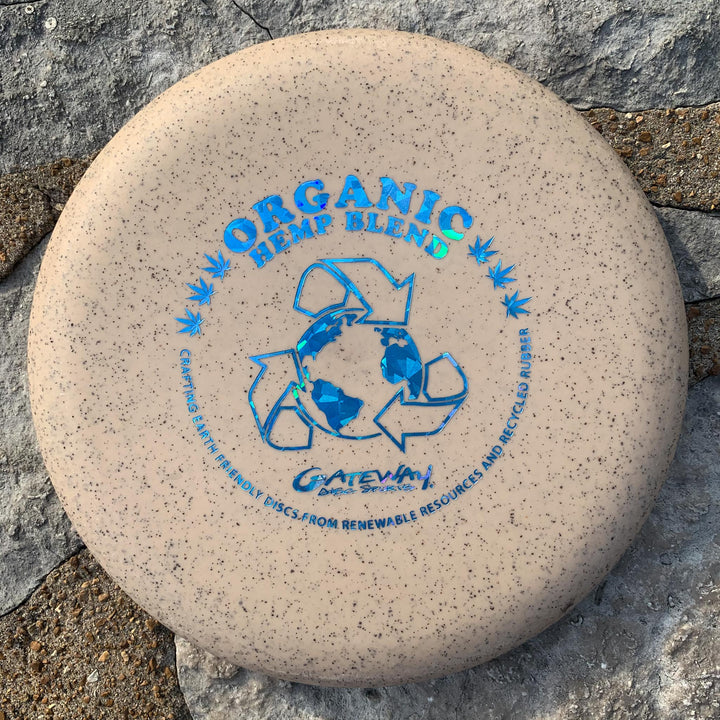 A beige Magic - Organic Hemp (H-SSS) frisbee with blue —Organic Hemp Blend—� text and recycling/earth graphics, designed for beginners with straight-flying performance, rests on a stone surface.