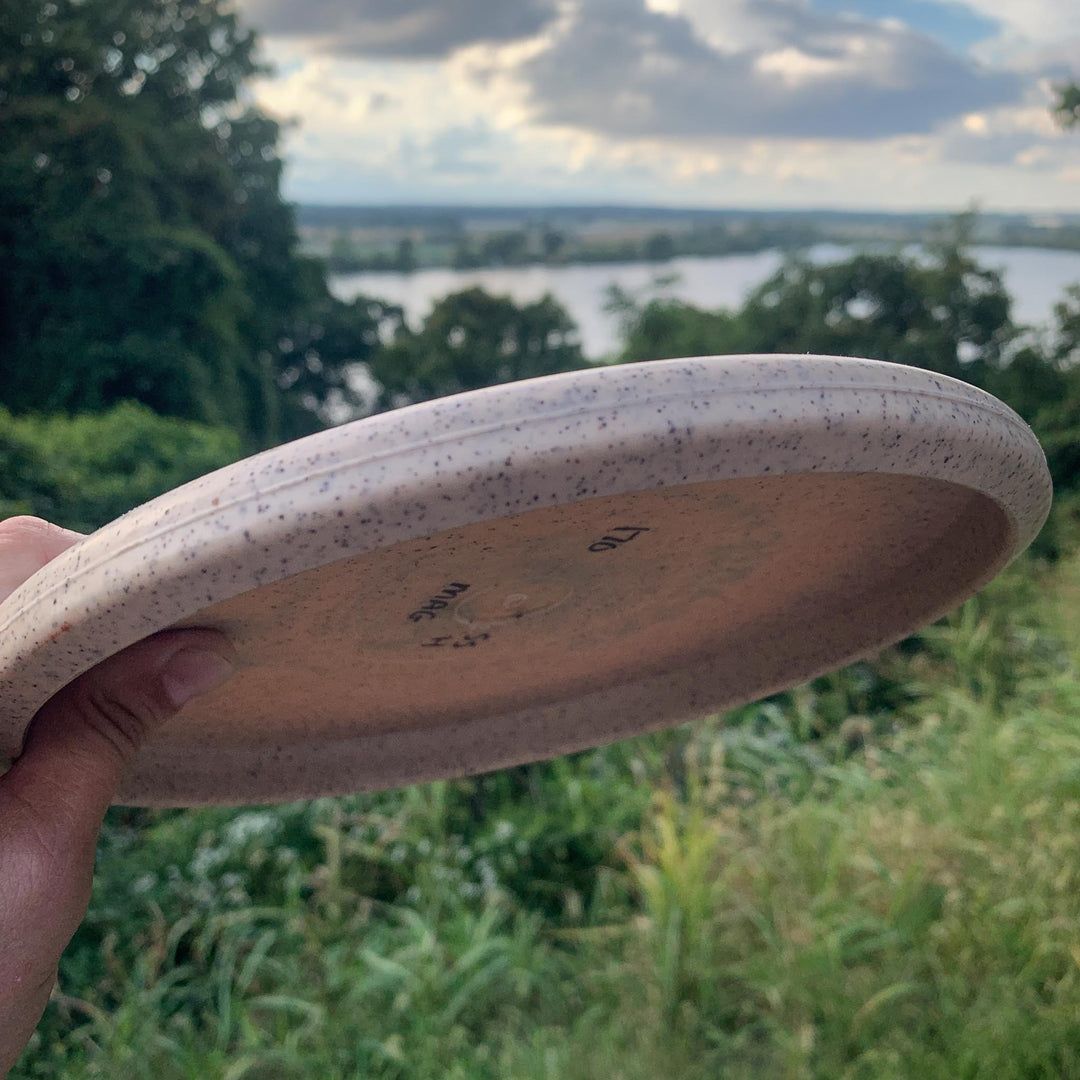 A hand holds the Magic - Organic Hemp (H-SSS), a speckled, beginner-friendly disc golf disc, outdoors with trees, grass, and a river under a cloudy sky.