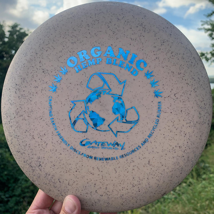 A hand holds the Magic - Organic Hemp (H-SSS) frisbee, labeled "Organic Hemp Blend," with blue recycling symbols and text about renewable resources and recycled rubber—a straight-flying, beginner-friendly disc.