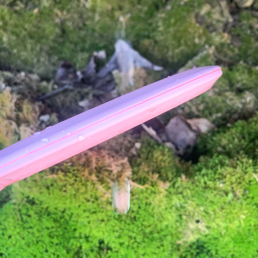The Mystic - SureGrip, a pink plastic disc known for its impressive glide and controllable mid-range, rests atop green moss and brown earth, highlighted by a single water droplet.