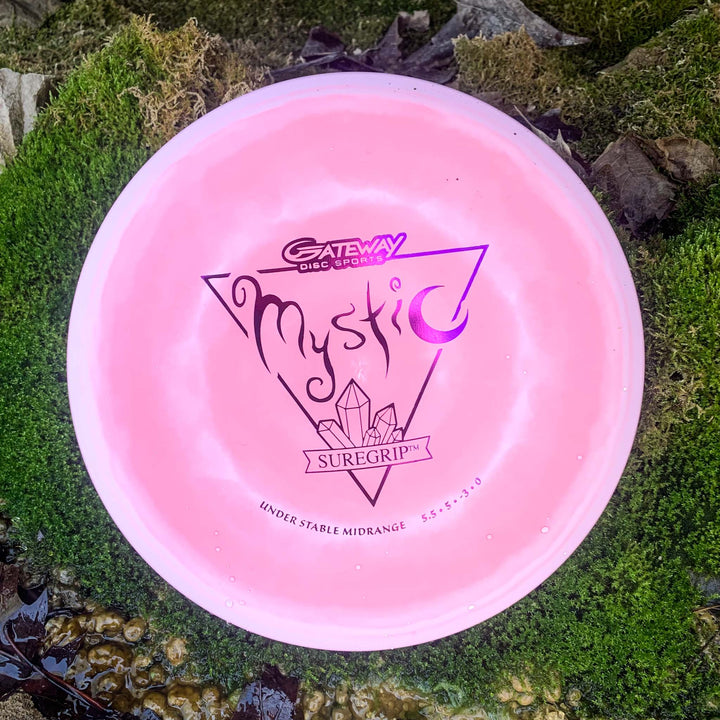 A pink Mystic - SureGrip disc from Gateway Disc Sports rests on green moss and rocks, showing its logo—ideal for disc golfers seeking a controllable mid-range with reliable, straight flight.