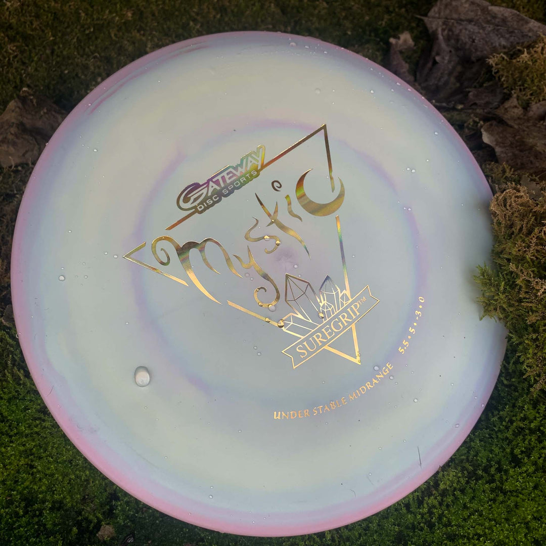 A Gateway Disc Sports Mystic - SureGrip controllable mid-range disc with a pastel swirl design and gold lettering rests on grass and moss, ideal for players wanting straight flight and excellent glide.