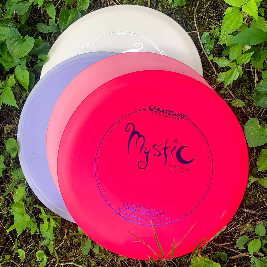 Three stacked disc golf discs—red, purple, and white—lie on grass amid green leaves. The top disc, the Mystic Lightweight-SureGrip™ by Gateway Disc Sports, offers controllable mid-range glide and effortless straight flight.