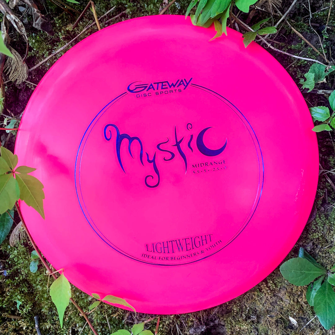 A vibrant pink Mystic Lightweight-SureGrip™ mid-range disc golf disc rests on mossy ground amid green leaves, ready for an effortless straight flight.