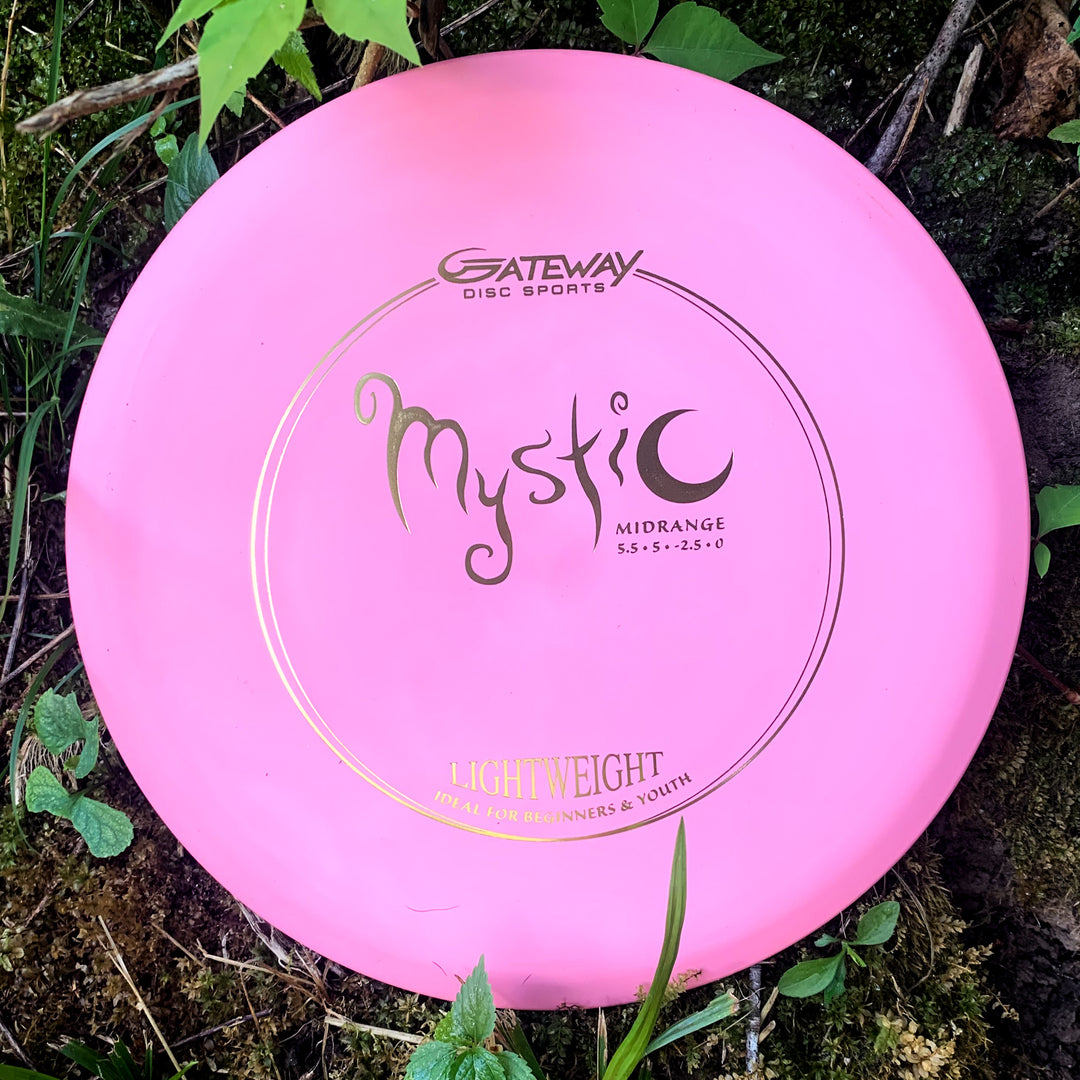A Mystic Lightweight-SureGrip™ mid-range disc golf disc by Gateway Disc Sports, in pink, known for straight flight and glide, is shown resting on grass and moss among plants. Labeled as lightweight.