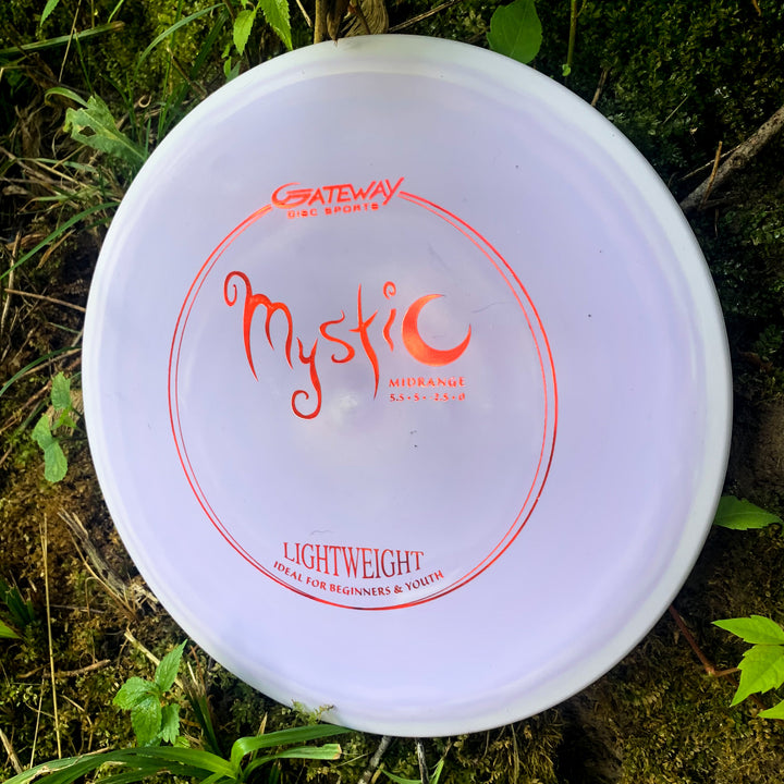 A white Mystic Lightweight- SureGrip™ disc golf disc with red text rests on grass—a reliable choice for effortless straight flights and precise mid-range throws.