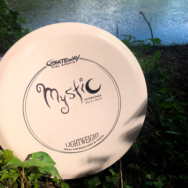 A white Mystic Lightweight- SureGrip™ mid-range disc golf disc lies by the water amid grass and foliage, ready to provide effortless straight flight.