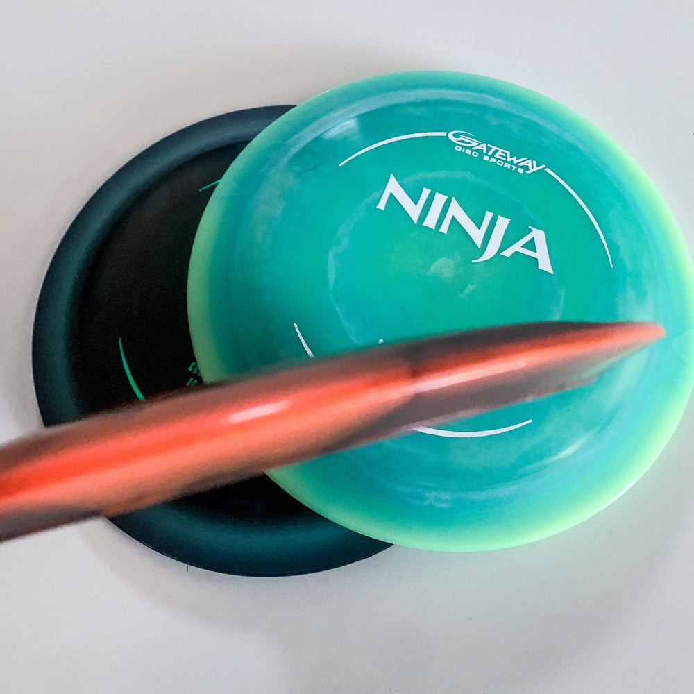 Two disc golf discs, including a green Ninja — (HD) Hyper Diamond featuring drag-reducing surface technology, and a dark-colored disc partially overlap on a white surface with a blurred red-tipped object in the foreground.