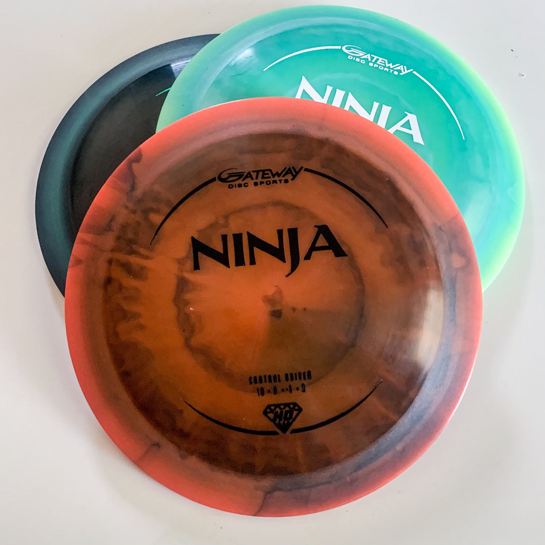 Three Ninja - (HD) Hyper Diamond disc golf discs in marbled red, green, and black are stacked on a white surface, each equipped with drag-reducing surface technology for enhanced performance.