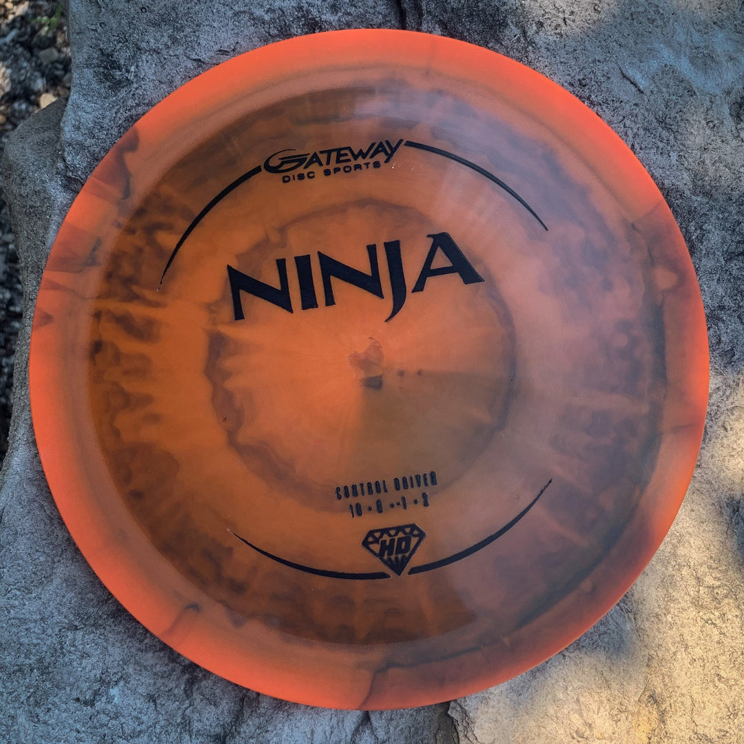 The orange and black Ninja - (HD) Hyper Diamond disc golf driver, with drag reducing surface technology, sits on a rough gray rock.