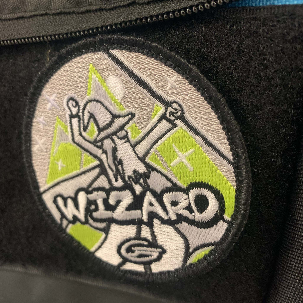 The Wizard Velcro Patch features a wizard with raised arms, hat, and beard, the word "WIZARD" beneath, and a green-gray background with stars and mountains—perfect for your Gateway Tour bag.