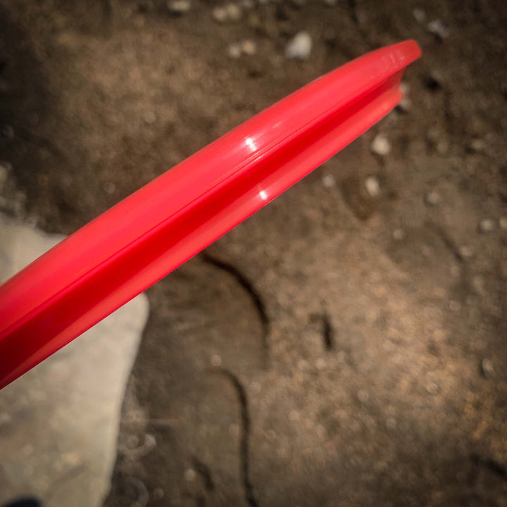 A close-up of the rim edge of a Prophecy - Diamond red plastic flying disc, held above a dirt ground background.