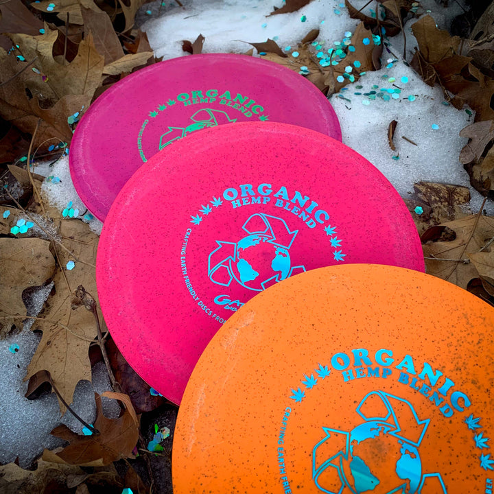 Three vibrant Prophecy - Suregrip Hemp disc golf discs rest on the ground among fallen leaves, snow patches, and blue confetti, showcasing their eco-friendly blend.