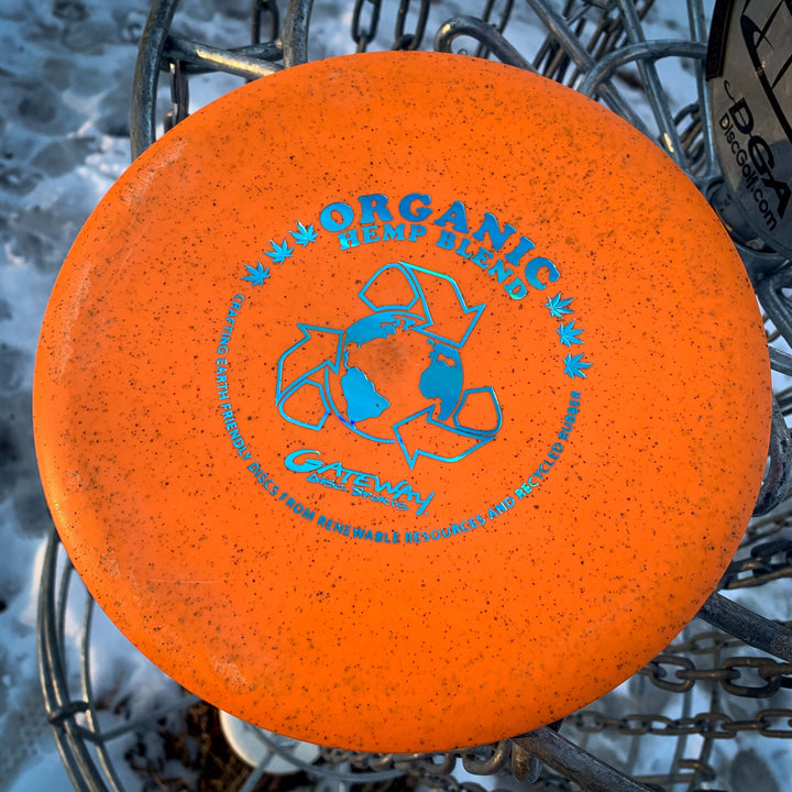 The Prophecy - Suregrip Hemp, an orange overstable mid-range disc with blue text and a recycling symbol, sits on a disc golf basket in snowy conditions—designed for consistent flight in any weather.