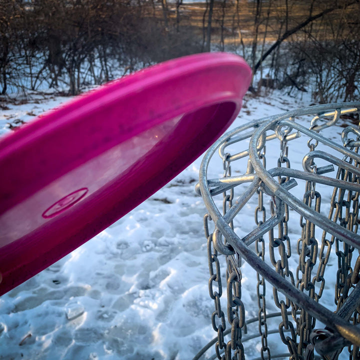 A close-up of a Prophecy - Suregrip Hemp disc about to be thrown into a metal disc golf basket on a snowy outdoor course.