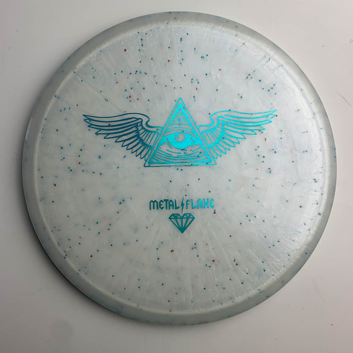 The Prophecy - Diamond Metal Flake is a white overstable mid-range disc golf disc with small colored flakes and a blue winged eye-in-triangle graphic, promising consistent flight.