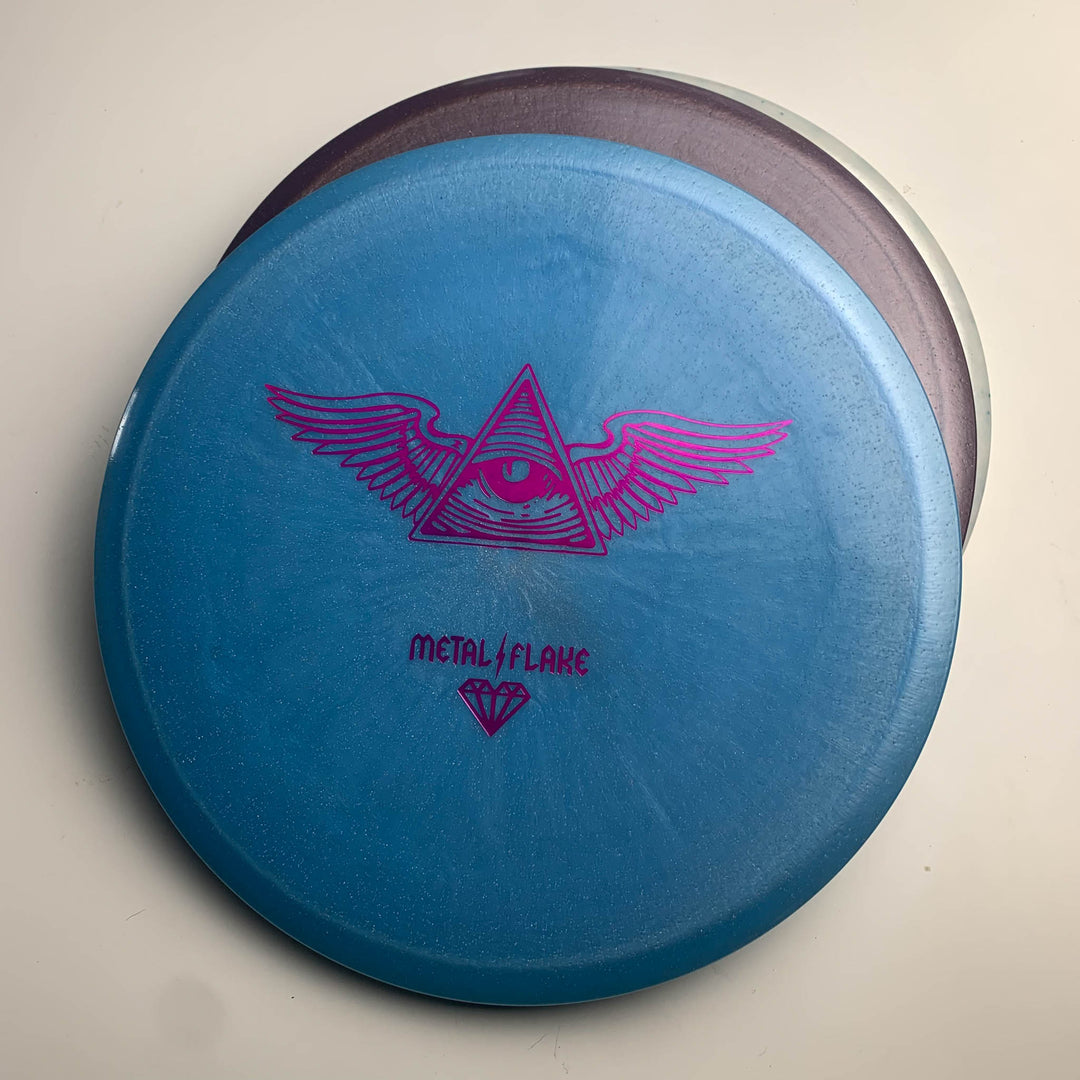 The Prophecy - Diamond Metal Flake disc features a blue premium Diamond blend, a pink winged eye pyramid design with "METAL/FLAKE", and delivers consistent flight on the course.