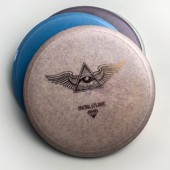 Three stacked Prophecy - Diamond Metal Flake discs; the top is light purple with metallic flecks, showing a winged all-seeing eye graphic and "Metal Flake" text—crafted in premium Diamond blend for reliable flight.