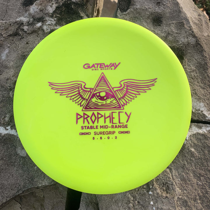 A yellow Gateway Prophecy - SureGrip stable mid-range disc with a winged triangle and eye design sits on large rocks, ready to provide a consistent flight.