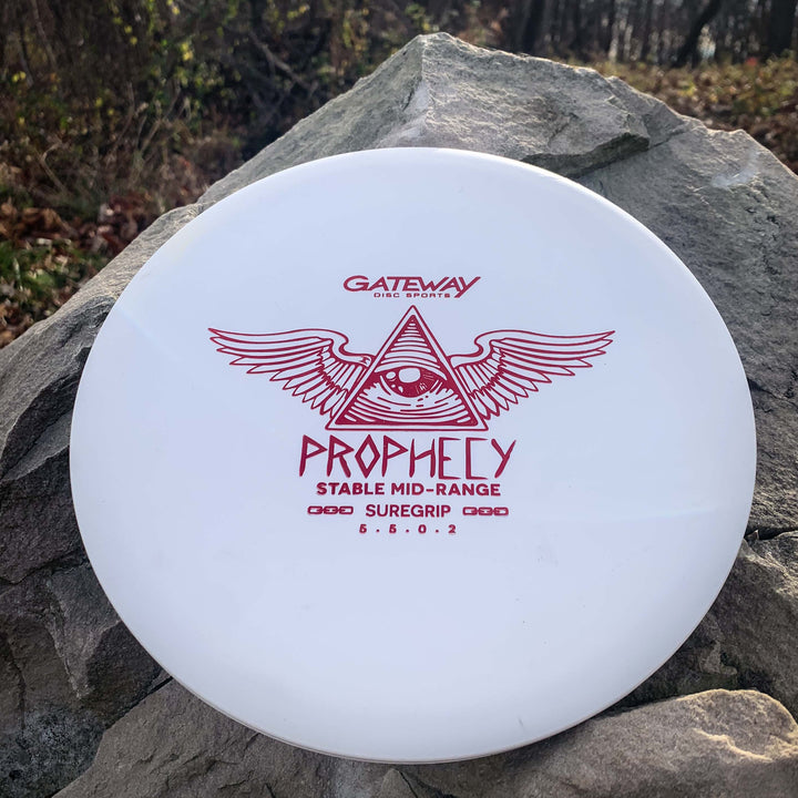 A white overstable mid-range disc golf disc labeled "Prophecy - SureGrip" with an eye and winged triangle graphic, featuring a grippy plastic blend and predictable fade, rests on a rock outdoors.