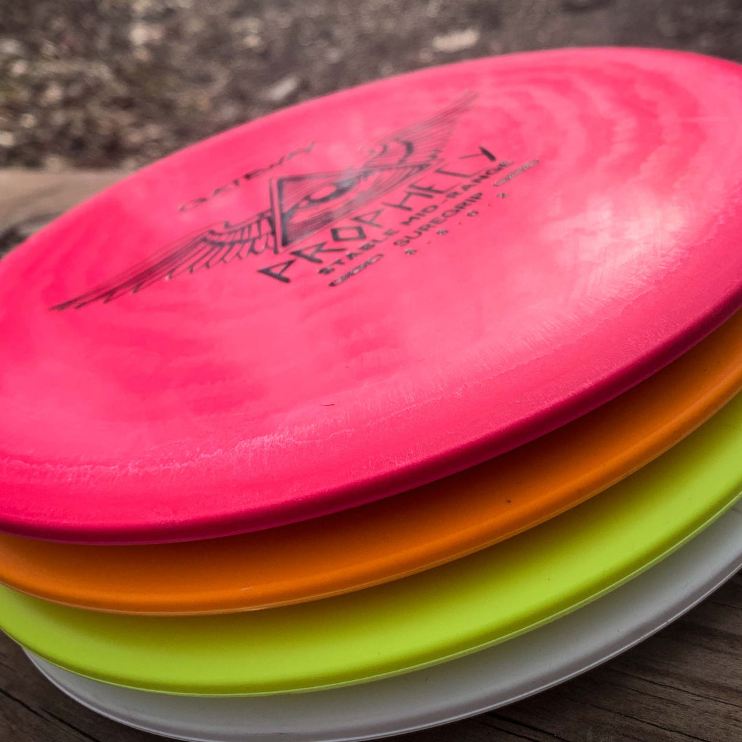 A stack of four colorful disc golf discs—pink, orange, yellow, and white—rests on a wooden surface, topped by the premium Prophecy - SureGrip mid-range disc known for its consistent flight.